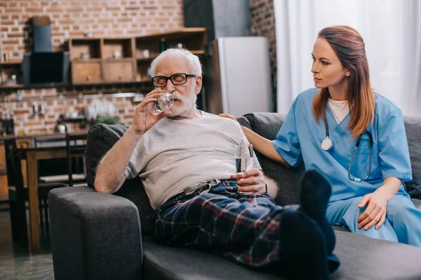 Hombre mayor tomando píldoras y enfermera sentado en el sofá - foto de stock