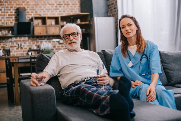 Старший пацієнт з таблетками і медсестрою сидить на дивані — стокове фото