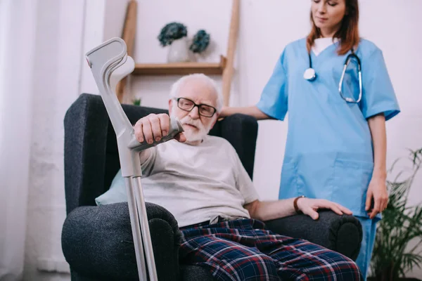 Hombre mayor con muleta sentado en silla por enfermera - foto de stock