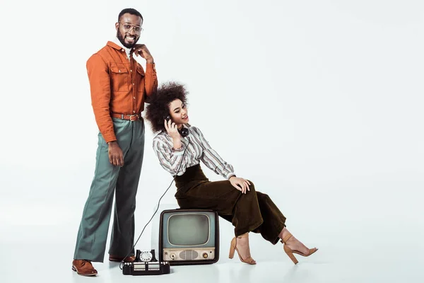 Happy african american retro styled girlfriend sitting on vintage television and and talking by phone on white — Stock Photo