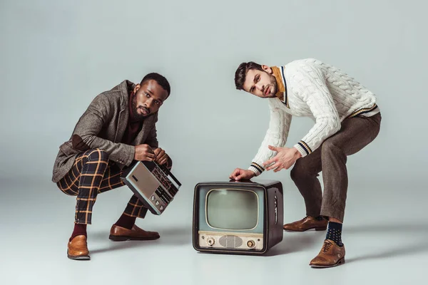 Amigos de estilo retro multicultural en cuclillas con radio vintage y televisión en gris - foto de stock