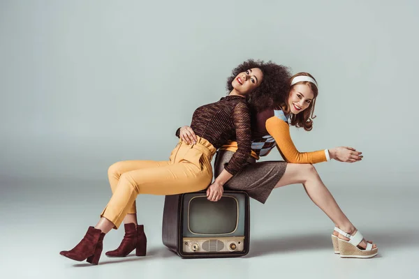 Happy multicultural retro styled girls posing on vintage television on grey — Stock Photo