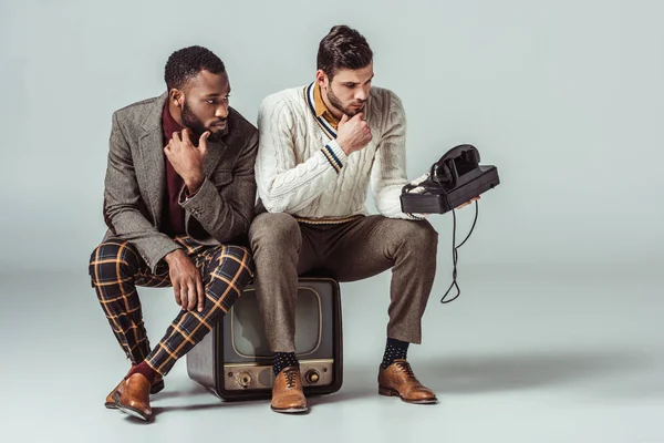 Multicultural retro styled friends sitting on vintage television and looking at stationary telephone — Stock Photo