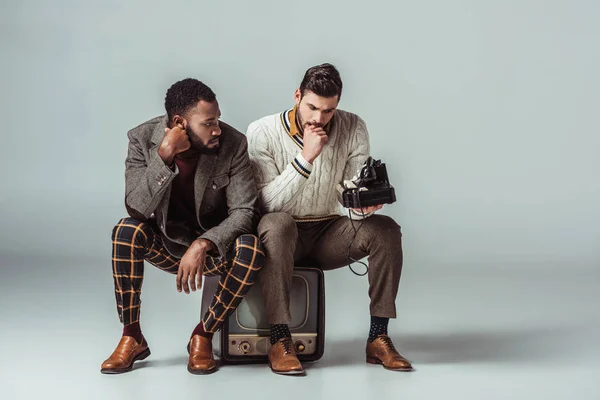 Multicultural retro styled friends sitting on vintage television and holding stationary telephone — Stock Photo