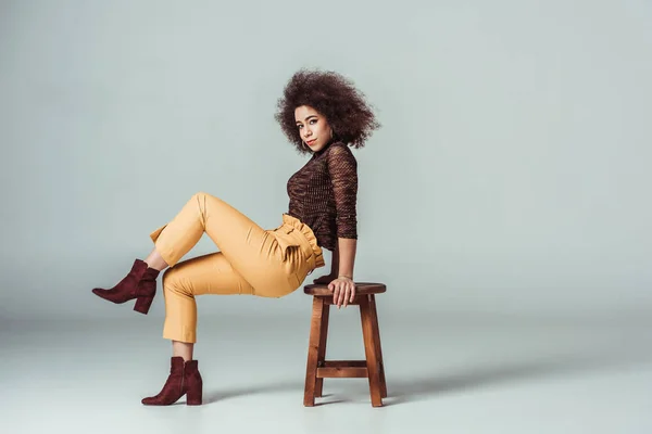 Vista lateral de mujer afroamericana en ropa retro apoyada en silla en gris - foto de stock