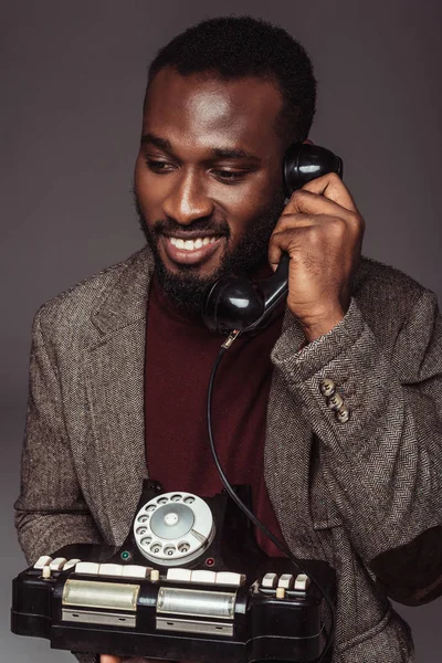 Lächelnd afrikanisch-amerikanischer Mann im Retro-Stil spricht mit einem alten stationären Telefon isoliert auf grau — Stockfoto