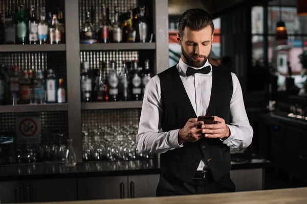 Camarero guapo usando teléfono inteligente en el mostrador de bar - foto de stock