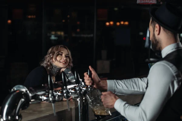 Barman vertiendo cerveza de grifos de cerveza en el vaso y mirando al visitante femenino - foto de stock