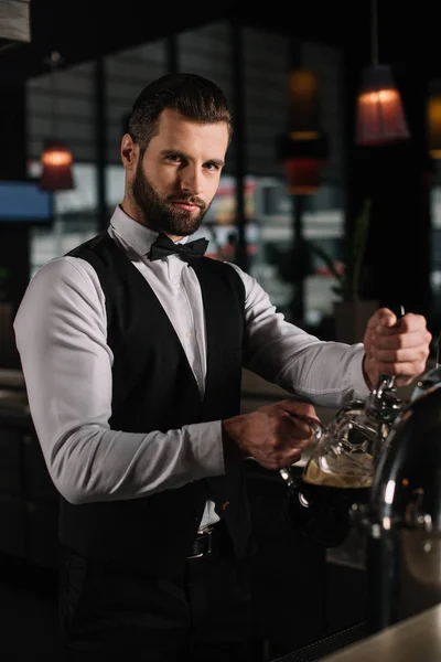 Handsome bartender pouring beer from beer taps into glass and looking at camera — Stock Photo