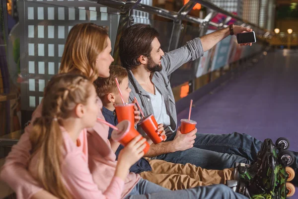 Vista lateral da família com bebidas tomando selfie enquanto descansa depois de patinar na pista de patinação — Fotografia de Stock