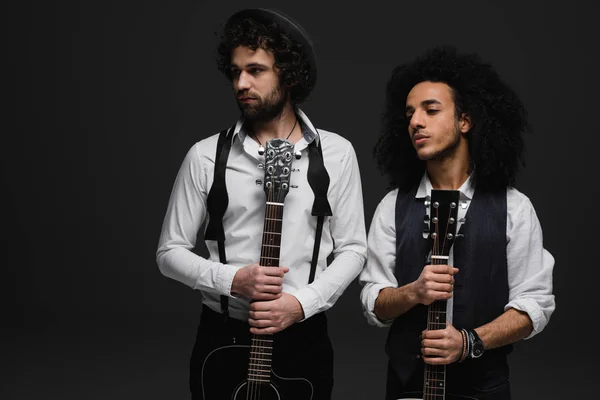 Dúo de jóvenes músicos guapos con guitarras acústicas mirando hacia otro lado en negro - foto de stock