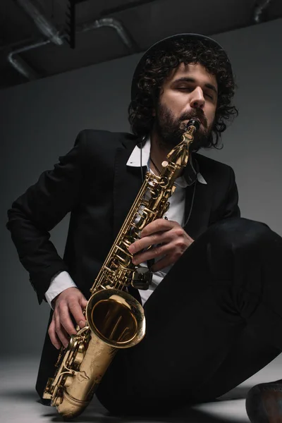 Músico artístico tocando el saxofón mientras está sentado en el suelo - foto de stock
