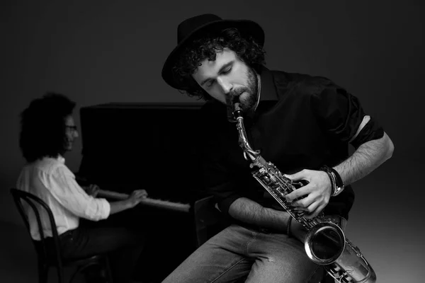 Black and white shot of man playing saxophone while his partner playing piano blurred on background — Stock Photo