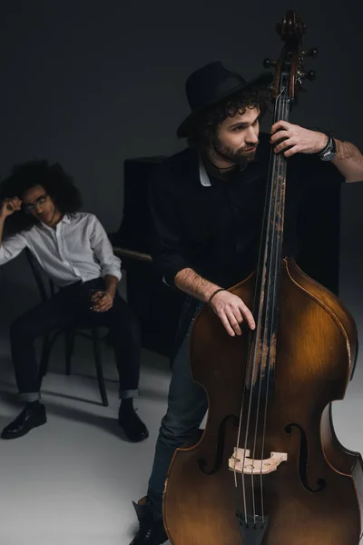 Hombre tocando el violonchelo mientras su compañero sentado al piano con un vaso de alcohol borroso en el fondo - foto de stock