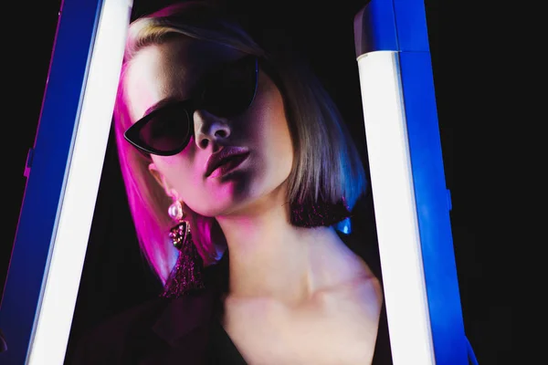 Стильная привлекательная девушка позирует в солнечных очках с двумя ультрафиолетовыми лампами для модной съемки, изолированная на черном — стоковое фото