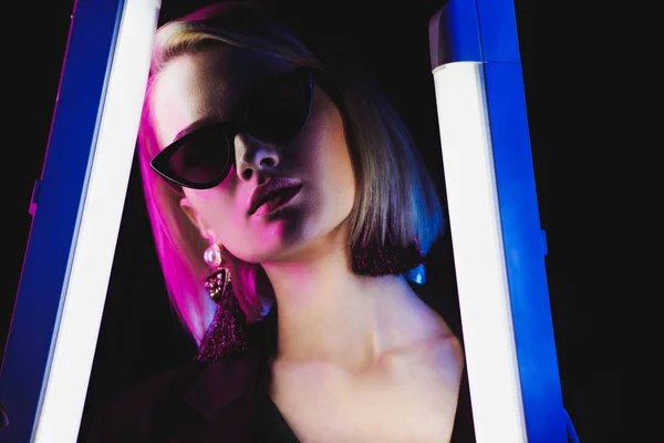 Elegante ragazza attraente posa in occhiali da sole con due lampade ultraviolette per le riprese di moda, isolato su nero — Stock Photo
