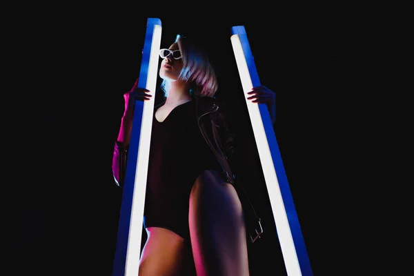 Стильная девушка позирует с двумя ультрафиолетовыми лампами для модной съемки, изолированная на черном — стоковое фото