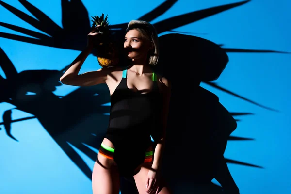 Fashionable girl posing in swimsuit with pineapple, on blue with palm leaves shadow — Stock Photo