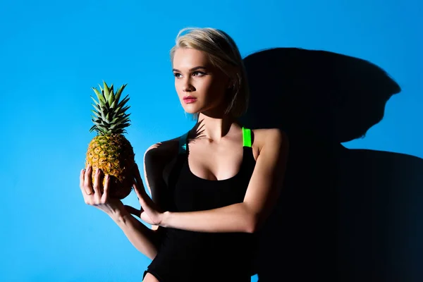 Beautiful slim girl posing in swimsuit with pineapple, on blue — Stock Photo