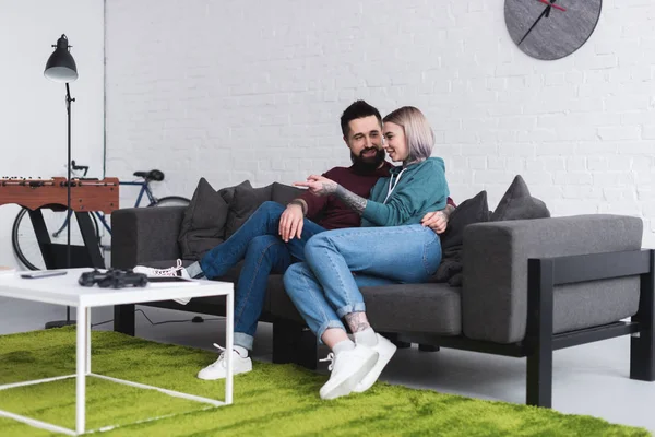 Lächelndes tätowiertes Paar, das sich auf dem Sofa im Wohnzimmer umarmt — Stockfoto