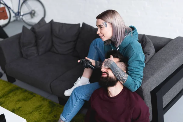 Tattooed girlfriend covering boyfriends eyes and playing video game — Stock Photo