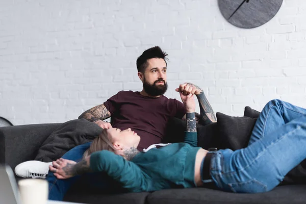 Pareja tatuada cogida de la mano en el sofá en la sala de estar - foto de stock