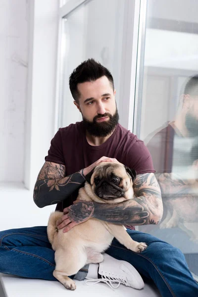 Tatuado bonito homem sentado no peitoril da janela e abraçando pug em casa — Fotografia de Stock