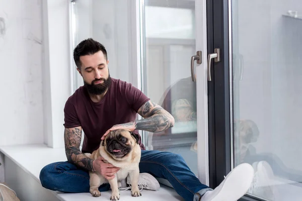 Hombre guapo tatuado sentado en el alféizar de la ventana y palming perro en casa - foto de stock