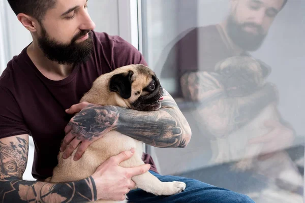 Tatuado homem sentado no peitoril da janela e abraçando pug em casa — Fotografia de Stock