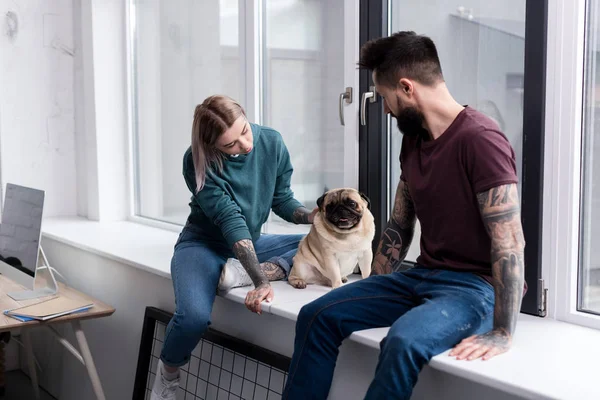 Pareja tatuada sentada en el alféizar de la ventana con pug en casa - foto de stock