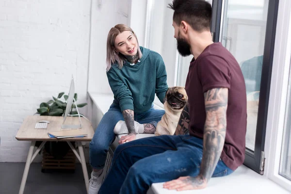 Feliz pareja tatuada sentada en el alféizar de la ventana con pug en casa - foto de stock