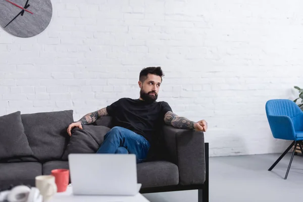 Hombre guapo tatuado sentado en el sofá y mirando a casa - foto de stock