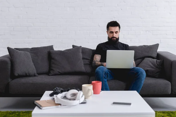 Hombre guapo tatuado usando el ordenador portátil en el sofá en casa - foto de stock