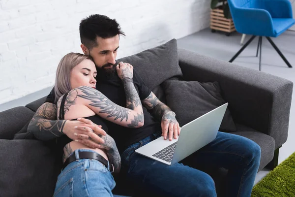 Pareja tatuada abrazando y mirando a la computadora portátil en casa - foto de stock