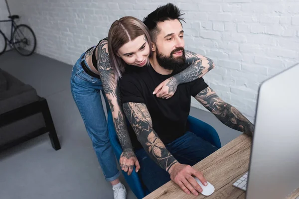 Татуированная подружка обнимает парня, пока он сидит дома за компьютером — стоковое фото