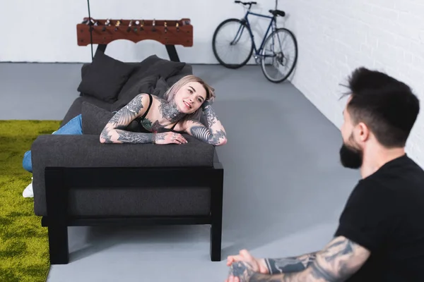 Smiling tattooed girlfriend lying on sofa and looking at boyfriend at home — Stock Photo