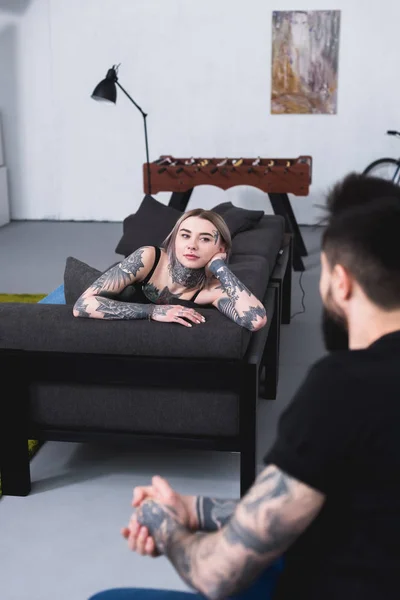 Tatuado namorada deitado no sofá e olhando para namorado em casa — Fotografia de Stock