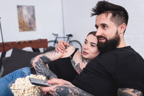 Affectionate tattooed couple watching tv with popcorn at home — Stock Photo