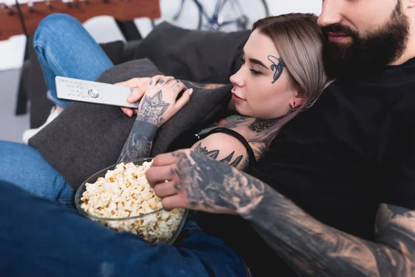 Imagen recortada de pareja tatuada viendo televisión con palomitas de maíz en casa - foto de stock