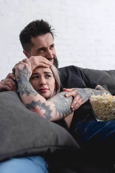 Asustada pareja tatuada viendo película de terror en el sofá en casa - foto de stock