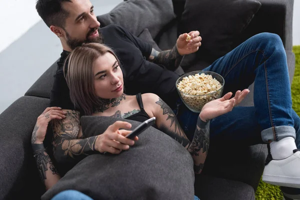 Pareja tatuada viendo la televisión en el sofá en casa - foto de stock