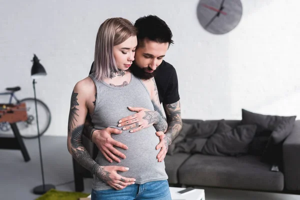 Feliz tatuado novio abrazando embarazada novia de nuevo en sala de estar - foto de stock