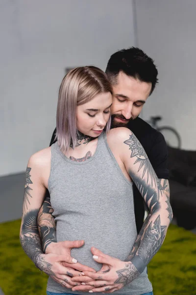 Щасливий татуйований хлопець обіймає вагітну дівчину ззаду вдома — стокове фото
