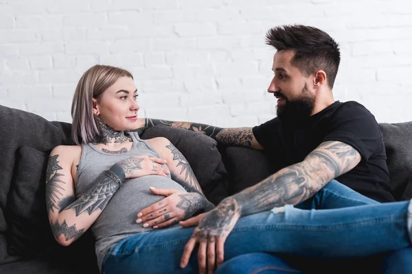 Tattooed pregnant girlfriend lying with boyfriend on sofa at home — Stock Photo