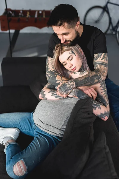 Alto ángulo vista de tatuado novio abrazando embarazada novia en sofá - foto de stock