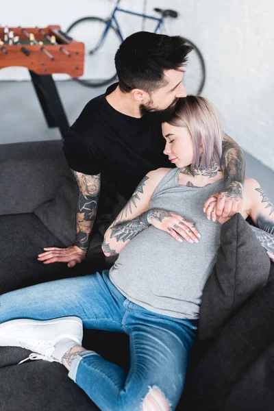 Vista de ángulo alto de novio tatuado abrazando a novia embarazada en el sofá en casa - foto de stock