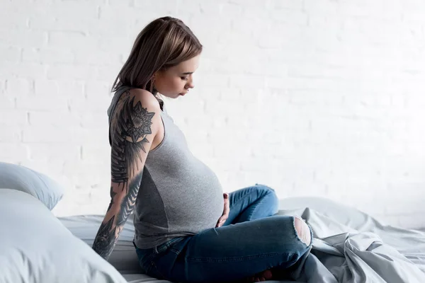 Side view of pregnant tattooed woman sitting on bed at home — Stock Photo