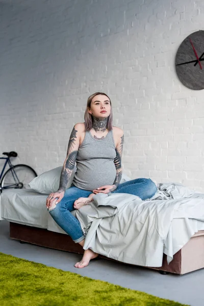 Mujer tatuada embarazada sentada en la cama en casa - foto de stock