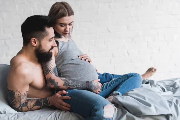 Novio abrazando embarazada tatuado novia en casa - foto de stock