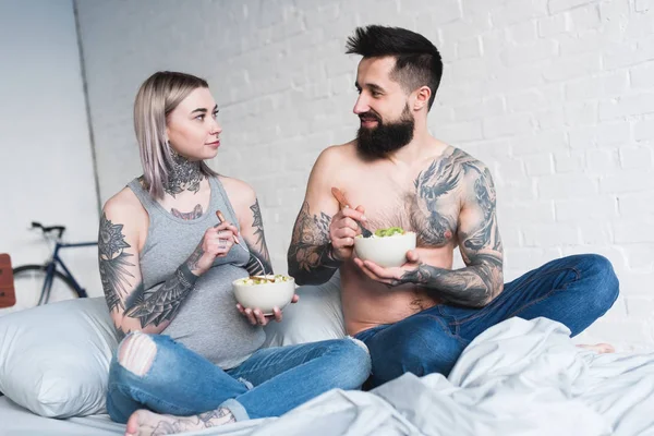 Pregnant tattooed girlfriend eating salad with boyfriend in bedroom — Stock Photo